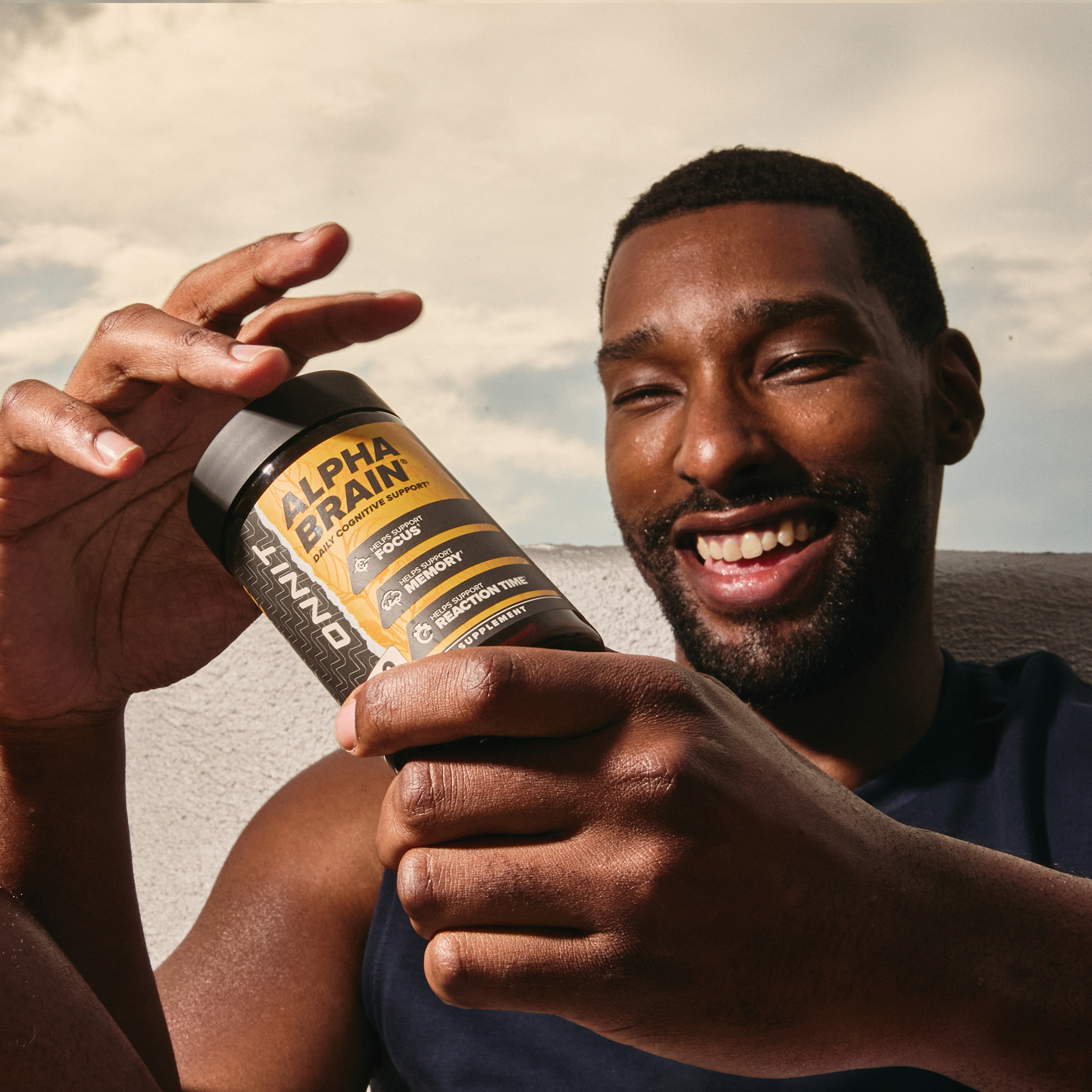 Alpha BRAIN supplement bottle with gold and black label held by a man outdoors.